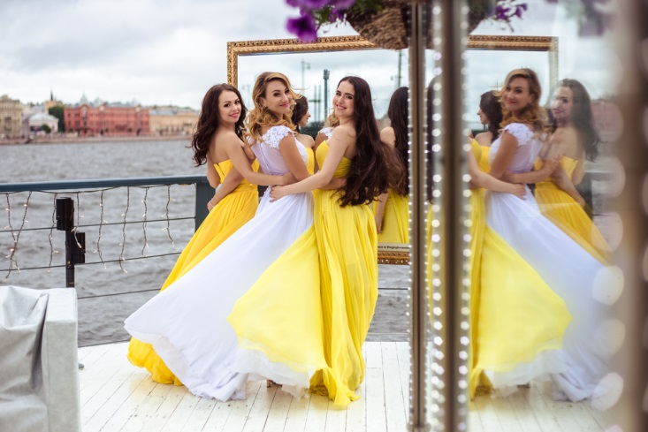Bride and two bridesmaids in yellow dresses smiling on dock at waterside wedding