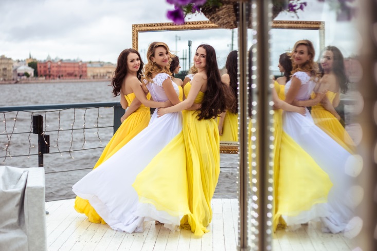 Bride and two bridesmaids in yellow dresses smiling on dock at waterside wedding